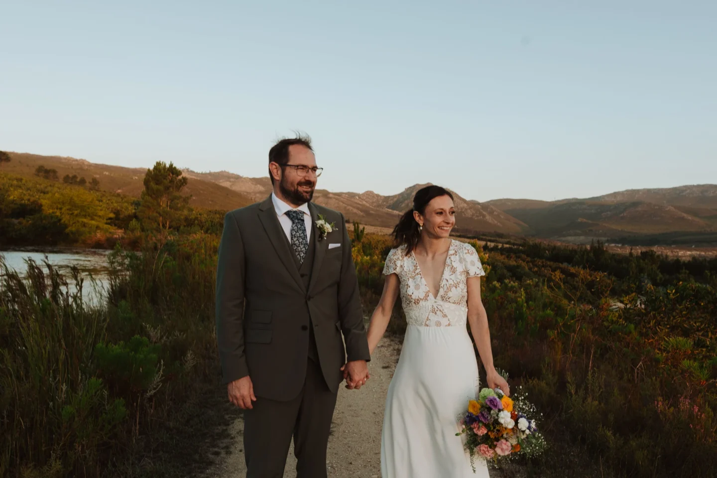 Bride and groom standing on the path beside the lake, hills behind them in the evening light, Old Mac Daddy, Elgin Valley