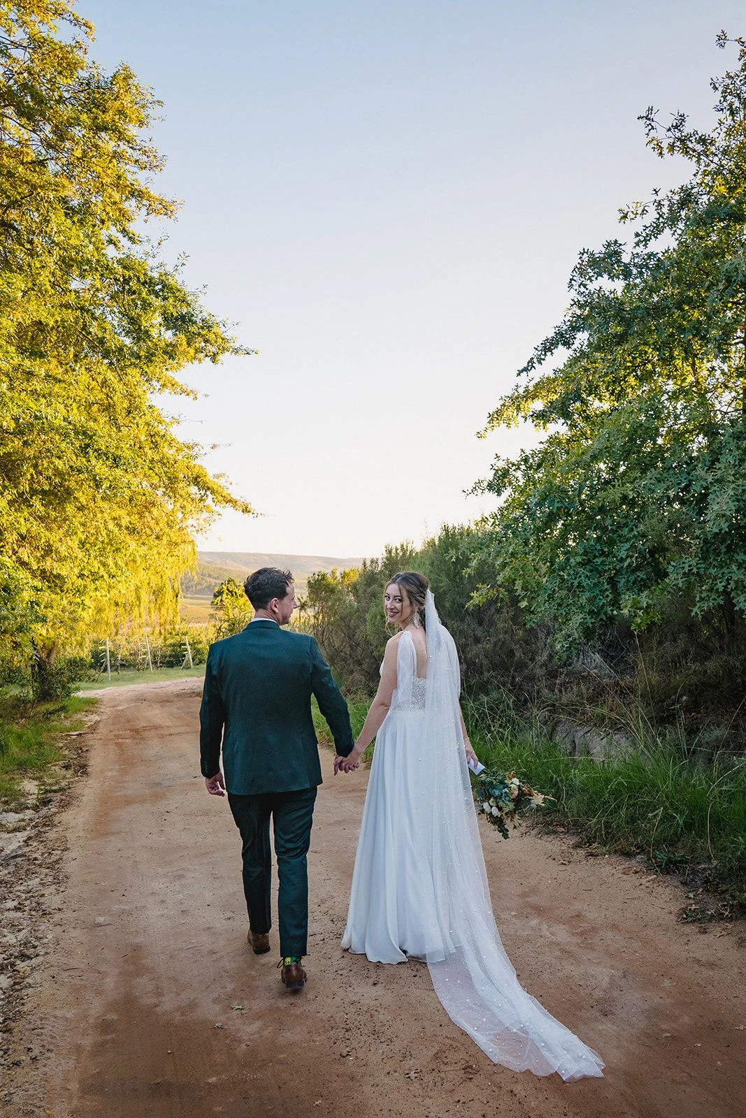 Bride and groom walking away along the farm road in the low sun, her veil trailing behind her, Old Mac Daddy, Elgin Valley