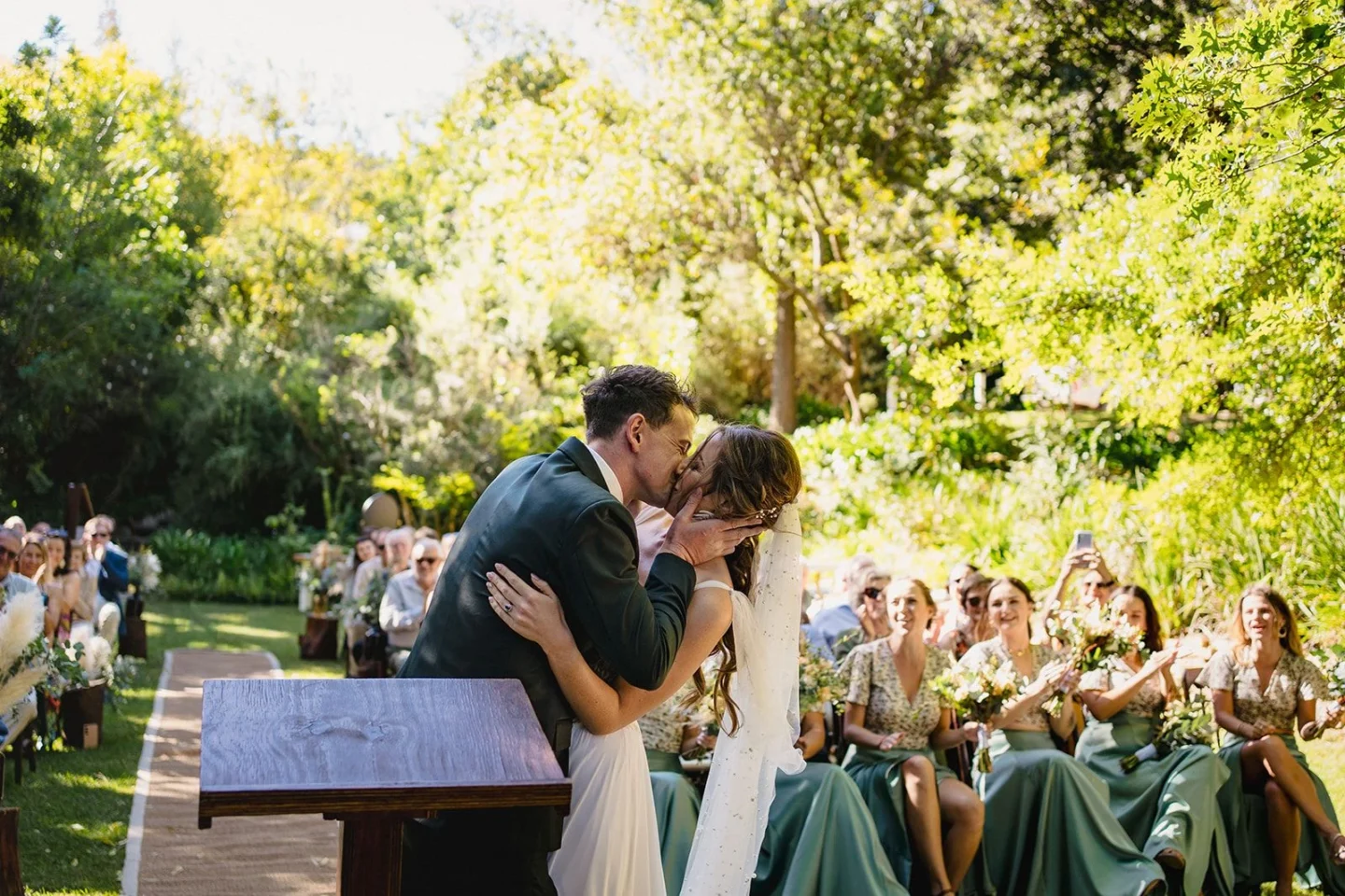 Bride and groom kissing at the ceremony while the bridal party in green cheers, Old Mac Daddy, Elgin Valley