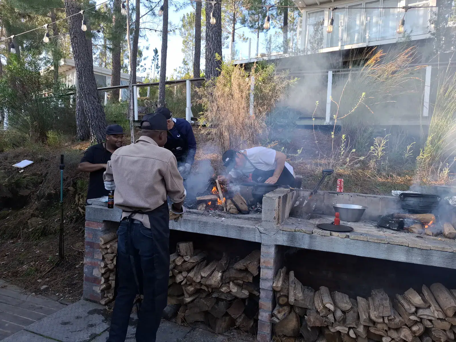 Home Teams cooking potjiekos over open fires at the long brick braai under the pines, firewood stacked below, Old Mac Daddy, Elgin Valley