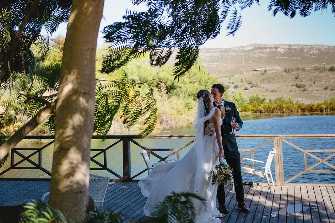 Bride and groom on the lakeside deck in the shade of a tree, hills across the water, Old Mac Daddy, Elgin Valley